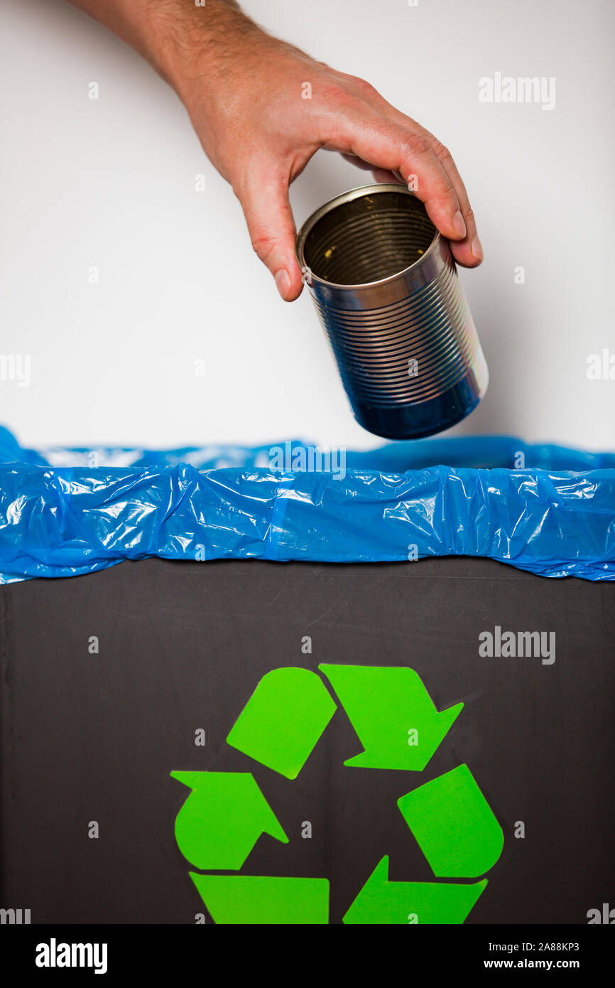 Hand putting tin can  into recycling bin. Person in a house kitchen separating waste. Black trash bin with blue bag and recycling symbol. Stock Photo