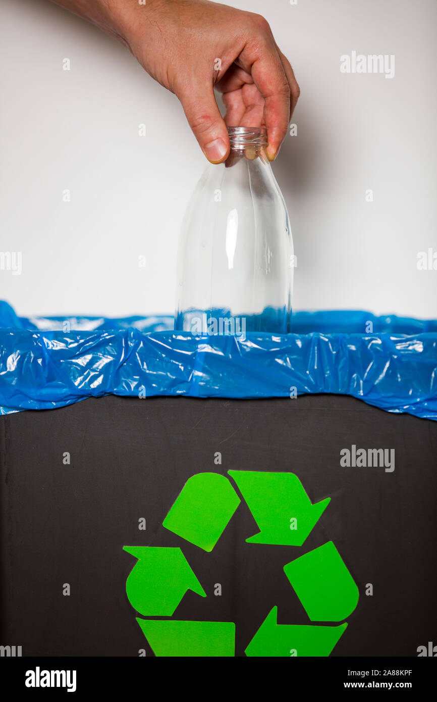 Hand putting glass bottle into recycling bin. Person in a house kitchen separating waste. Black trash bin with blue bag and recycling symbol. Stock Photo