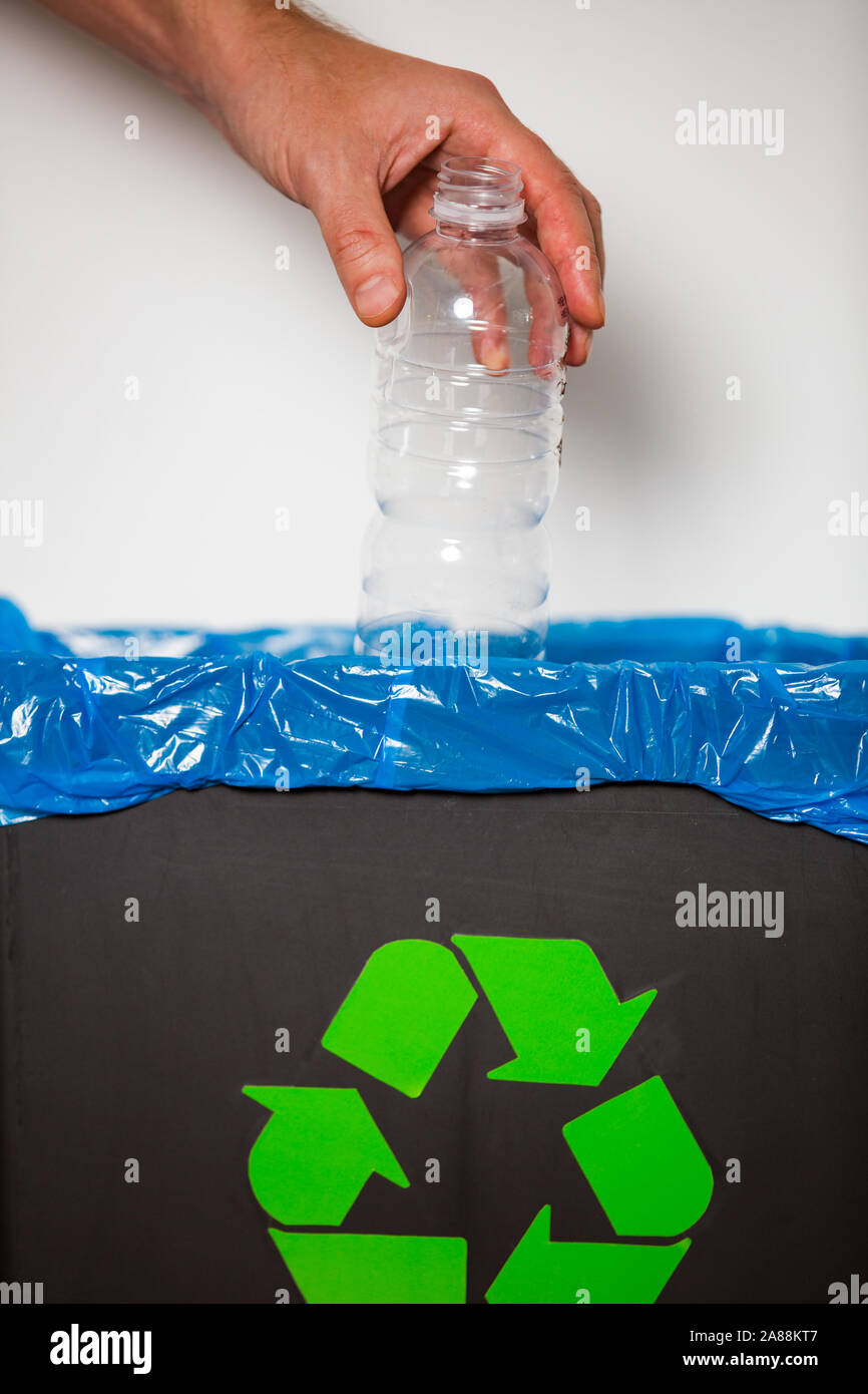 Hand putting single-use plastic bottle into recycling bin. Person in a house kitchen separating waste. Black trash bin with blue bag and recycling Stock Photo