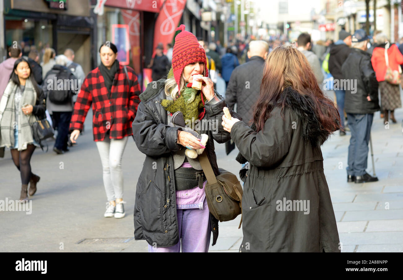 Two mature women eating in the street. One with red woolly hat. Stock Photo