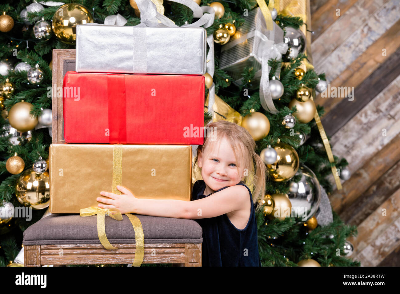 Portrait of elementary age girl wearing blue dress standing beside christmas tree holding big pile of wrapped gift boxes happy to get a lot of present Stock Photo