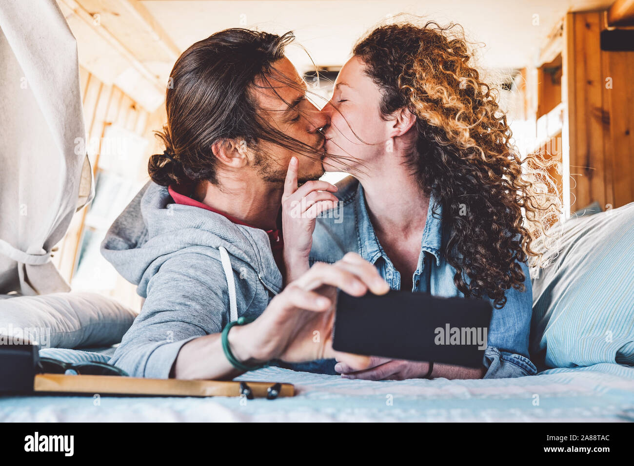 Happy couple kissing in vintage mini van during their roadtrip - Young romantic lovers having tender moments while taking photo with mobile smartphone Stock Photo