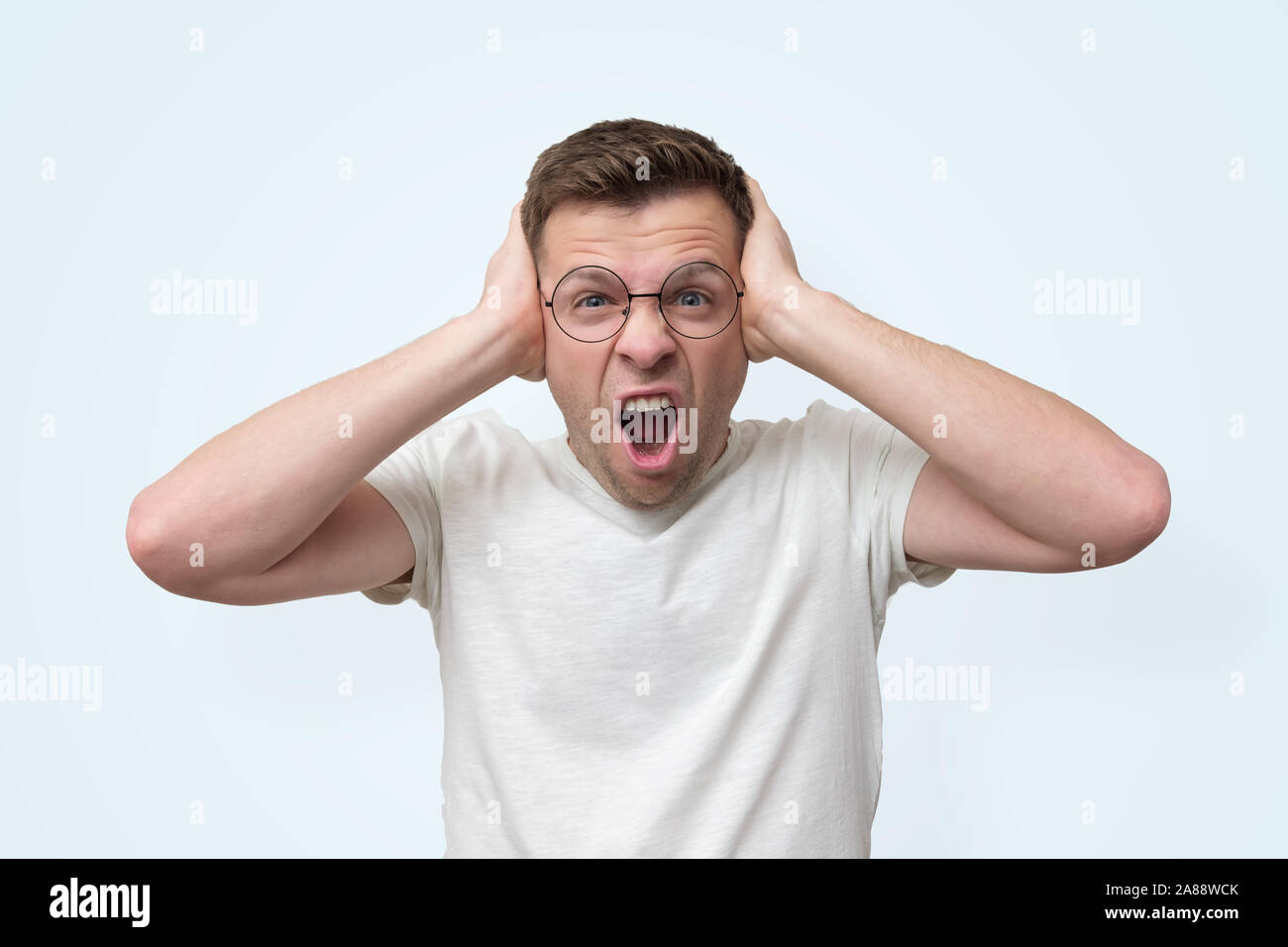 Annoyed young man in glasses screaming and covering his ears because he can not stand the noise. Negative facial human emotion Stock Photo