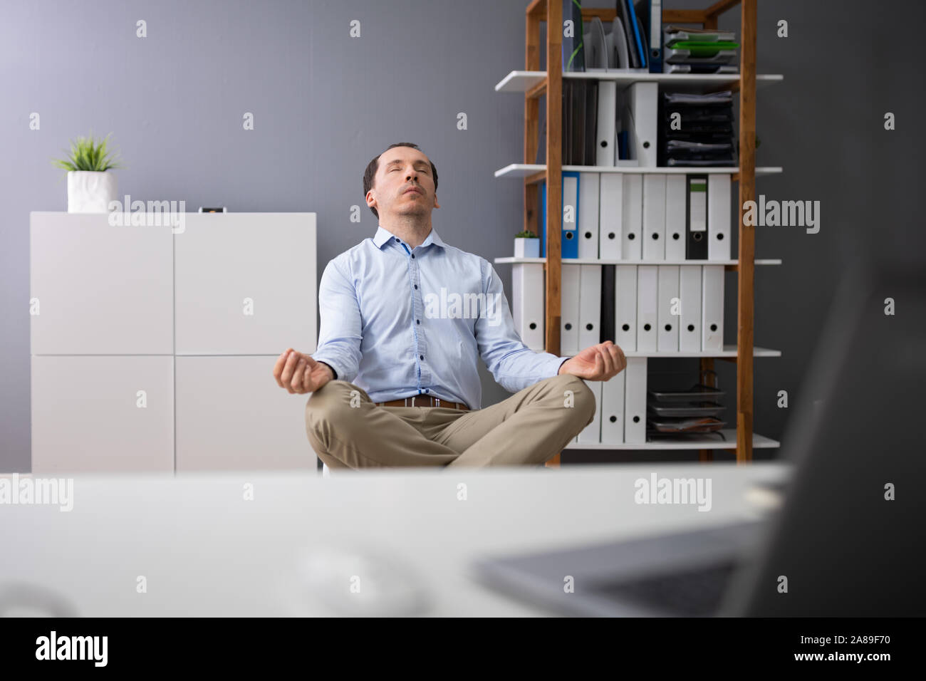 Young Businessman Doing Yoga Behind Desk In Office Stock Photo - Alamy