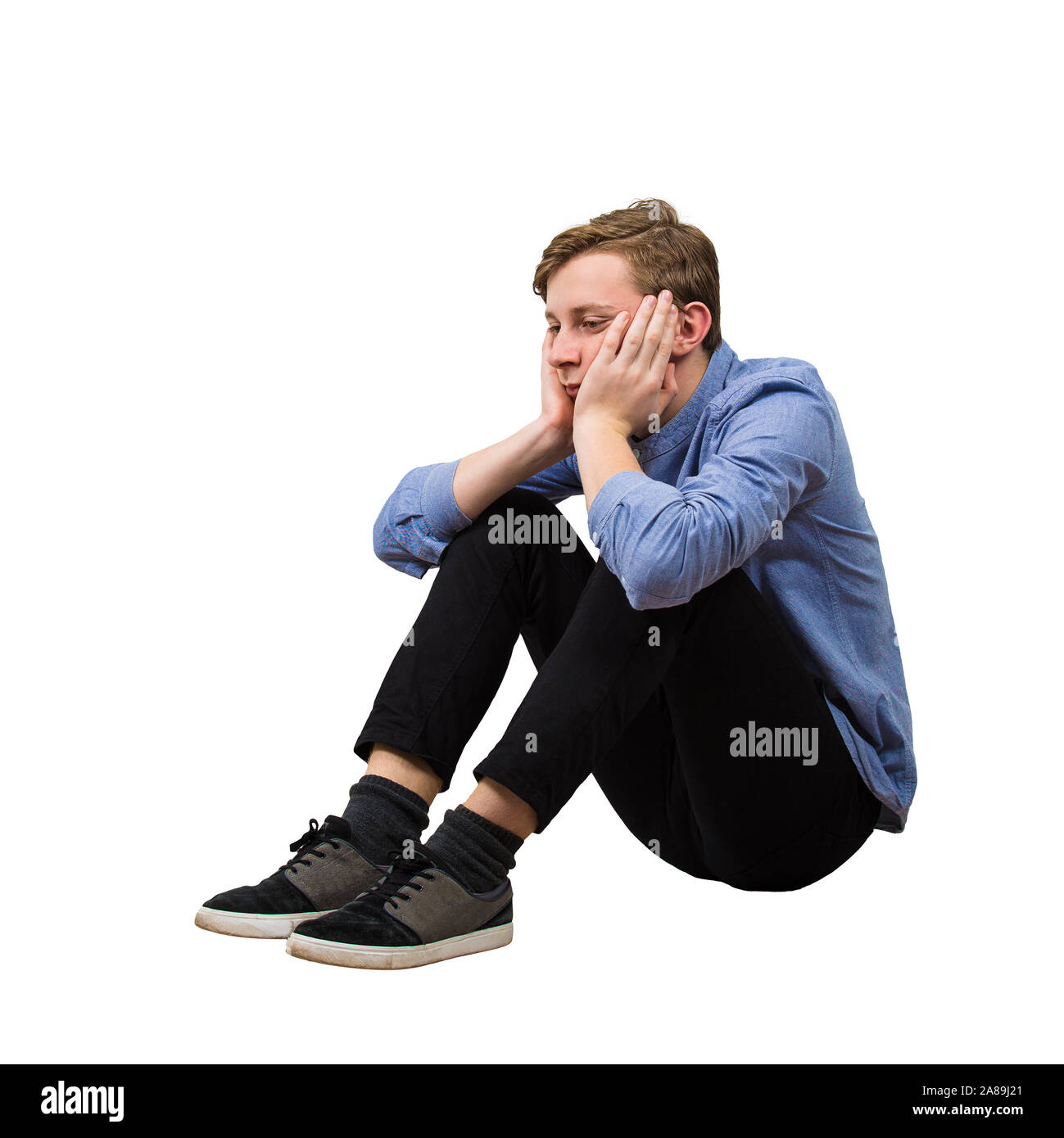 Full length side view of upset and tired boy teenager sitting on the floor keeps hands under chin looking down thoughtful isolated over white backgrou Stock Photo