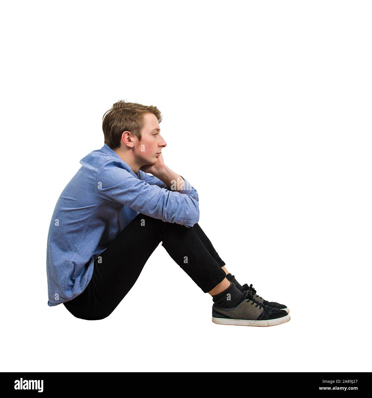Full length side view of upset and tired boy teenager sitting on the floor keeps hand under chin looking ahead thoughtful isolated over white backgrou Stock Photo