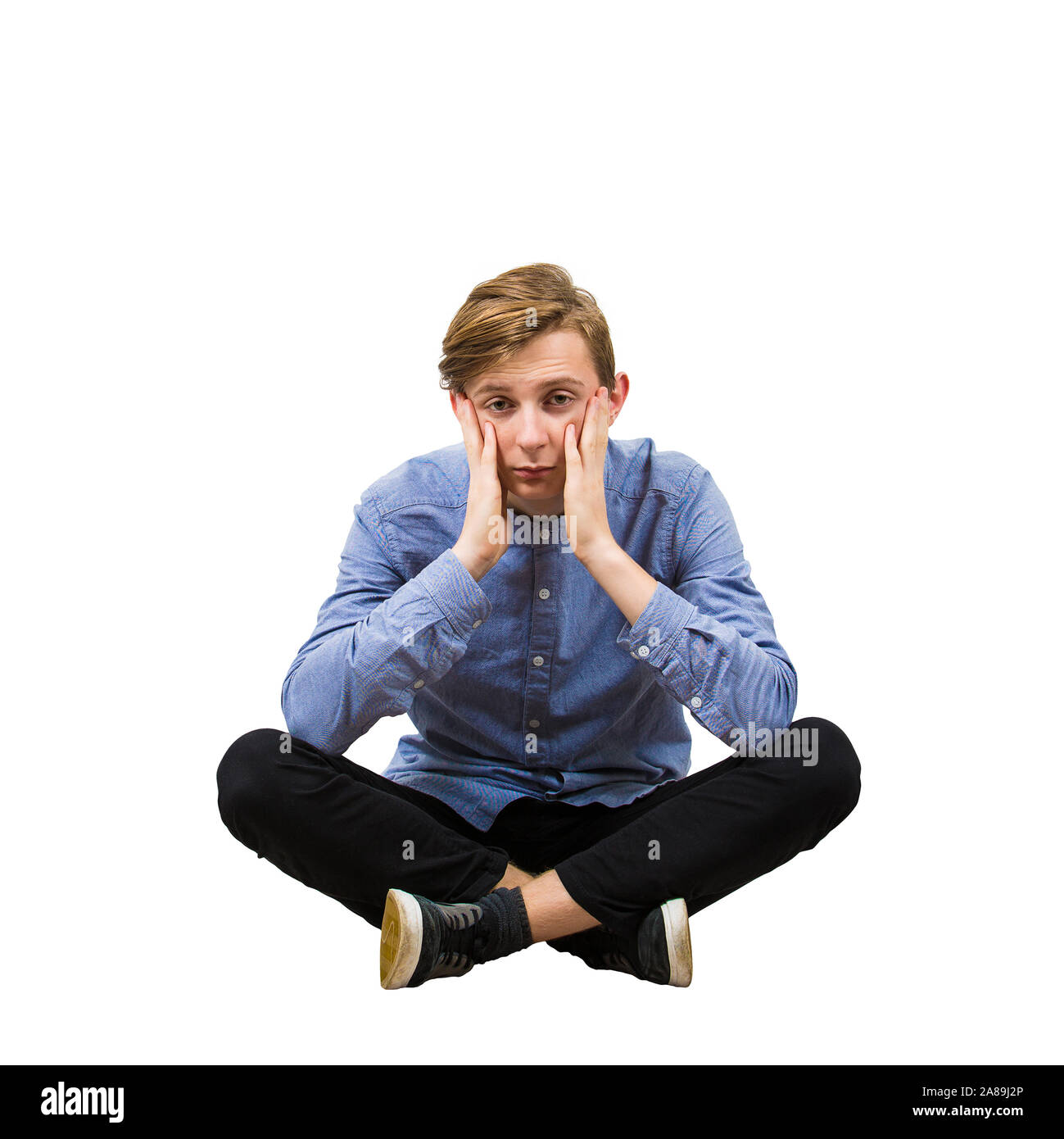 Upset and stressed teenage guy seated on the floor with crossed legs, covering his cheeks with both hands feel tired and sad isolated over white backg Stock Photo