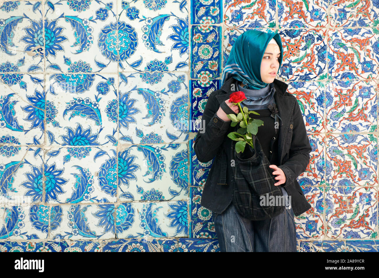 ISTANBUL - MAY 4, 2010: A young Turkish woman in bright headscarf stands holding a red rose against colorful patterned tile background. Stock Photo