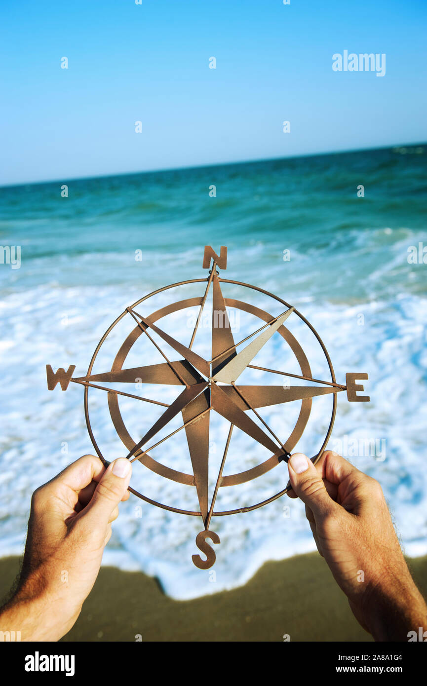 Hands holding simple metal compass rose in front of shoreline with crashing waves Stock Photo
