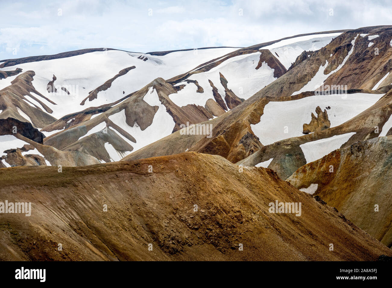 Snow-capped undulating barren hills of lava are set below a bright cloudy sky in Iceland Stock Photo