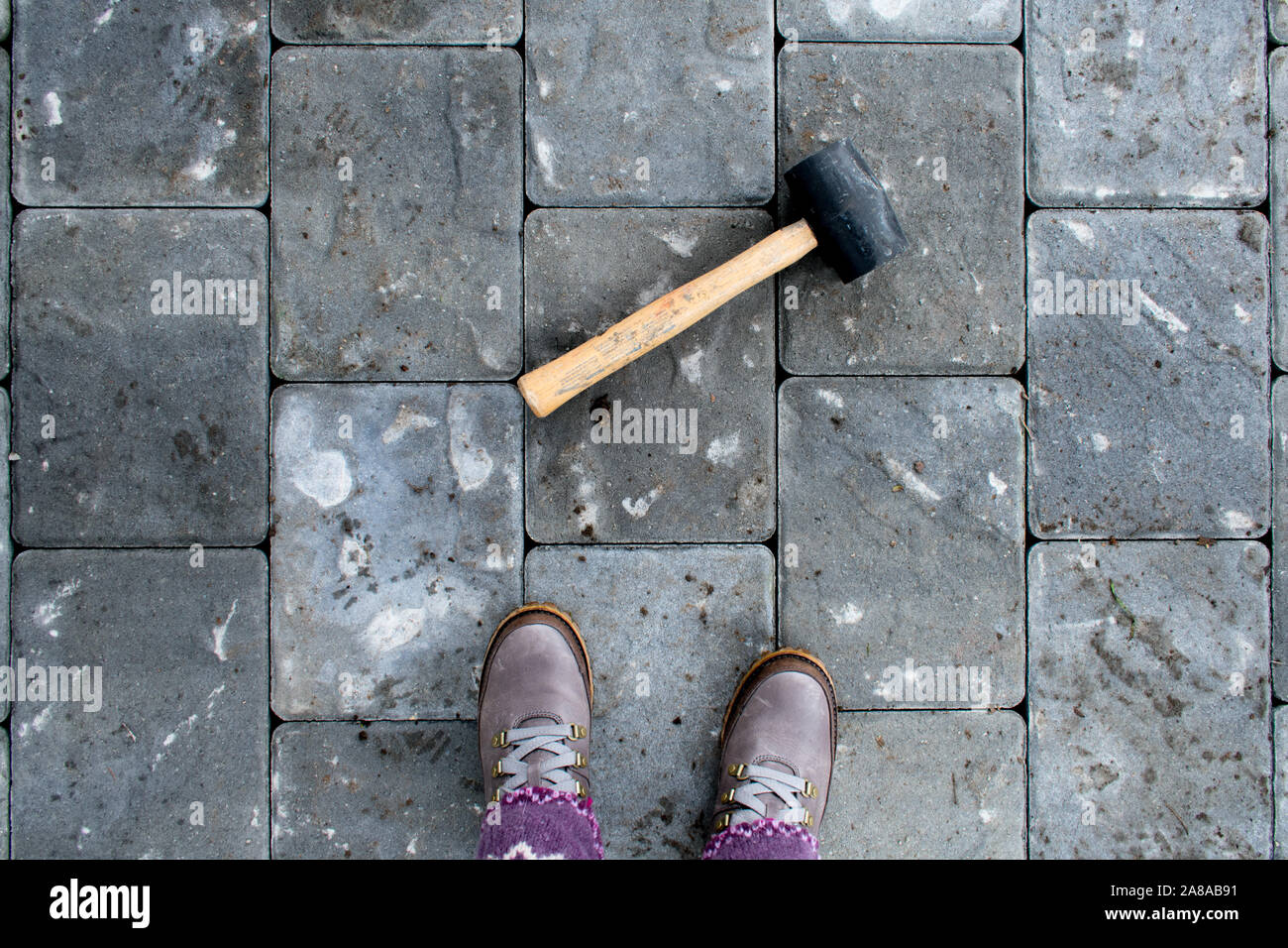 Woman in boots looking down at her feet and mallet on paver patio, over the shoulder view, MR Stock Photo