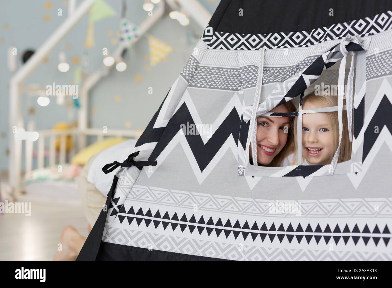 Happy mother with her 3 years old girl watching out of tent playhouse in playroom Stock Photo