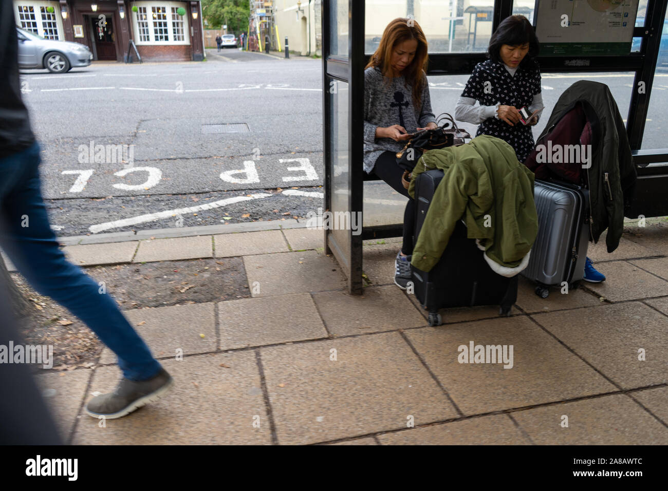 two asian women sitting at a bus stop with suitcases while people walk by Stock Photo