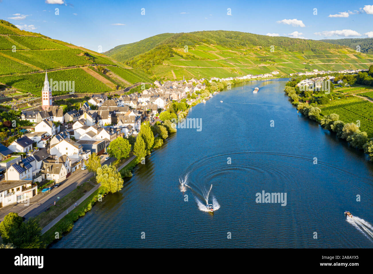 Hills with vineyards and church in Merl village of Zell (Mosel) town, Rhineland-Palatinate, Germany. Water skiing, barges, speedboats on the Moselle r Stock Photo