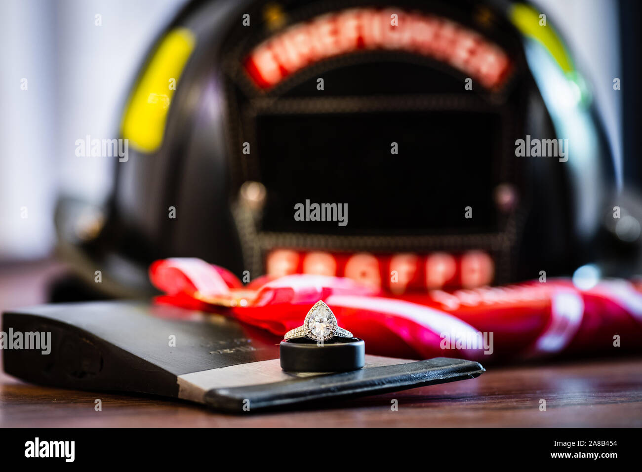 A teardrop, or pear, shaped diamond engagement ring on top of a wedding band and fireman's ax with a firefighter helmet blurred in the background. Stock Photo