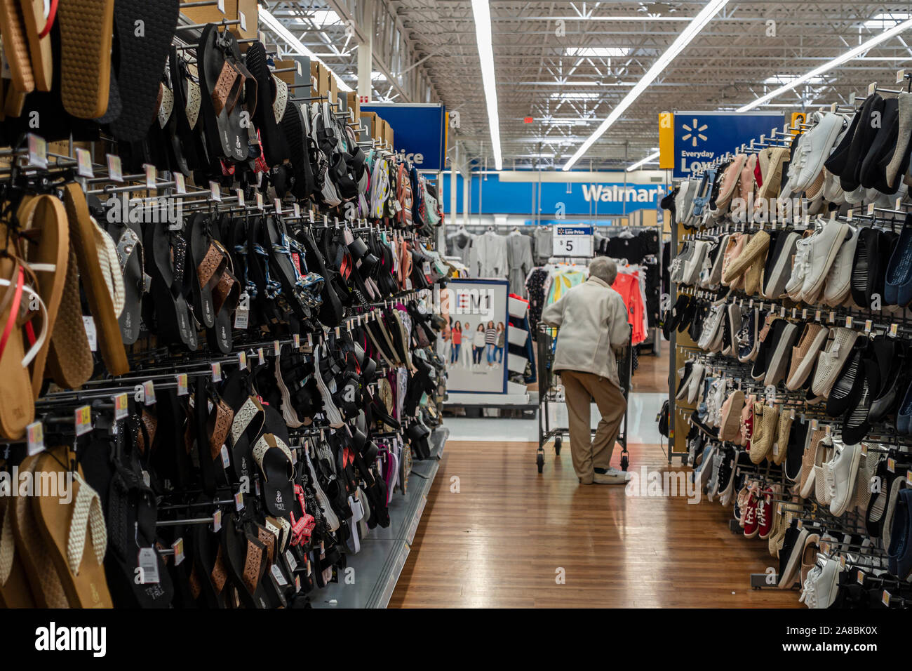 A gray-haired adult senior Caucasian woman holds on to a shopping cart while trying on shoes in Walmart. USA. Stock Photo