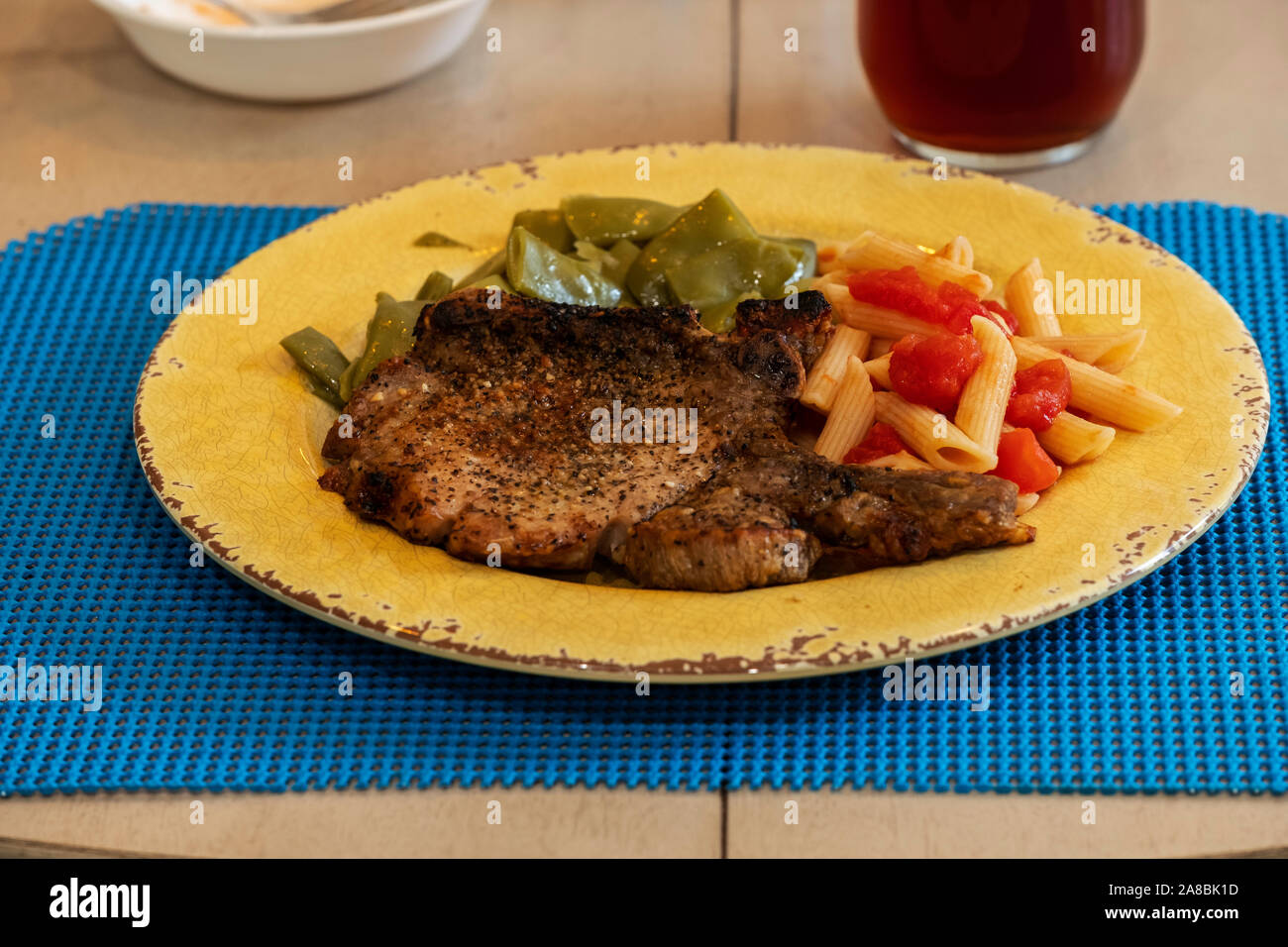 A grilled pork chop dinner with penne pasta and tomatoes, Italian green beans on a yellow plate. USA. Stock Photo