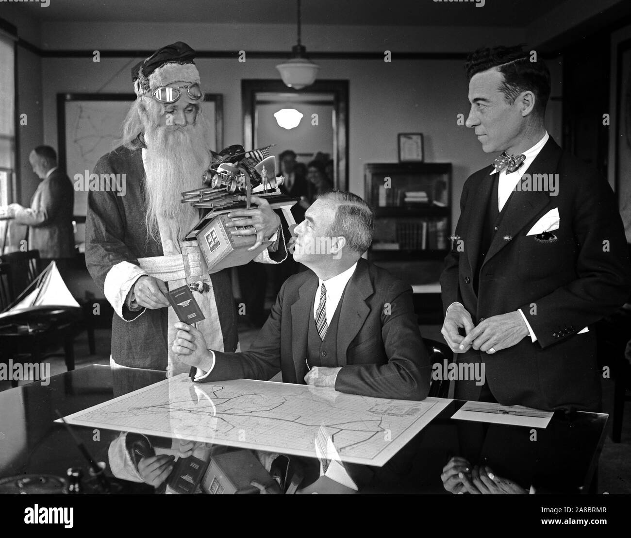 Santa Claus receives aeroplane pilot's license from Assistant Secretary of Commerce. Although there may not be sufficient snow for his reindeer sleigh, Santa Claus will still be able to deliver his load of presents on time this Christmas by using the air route. The old saint called at the Commerce Department in Washington today where he is shown receiving an aeroplane pilot's license from Assistant Secretary of Commerce. for Aeronautics William P. MacCracken, while Clarence M. Young (right) Director of Aeronautics, Department of Commerce, looks on. Stock Photo