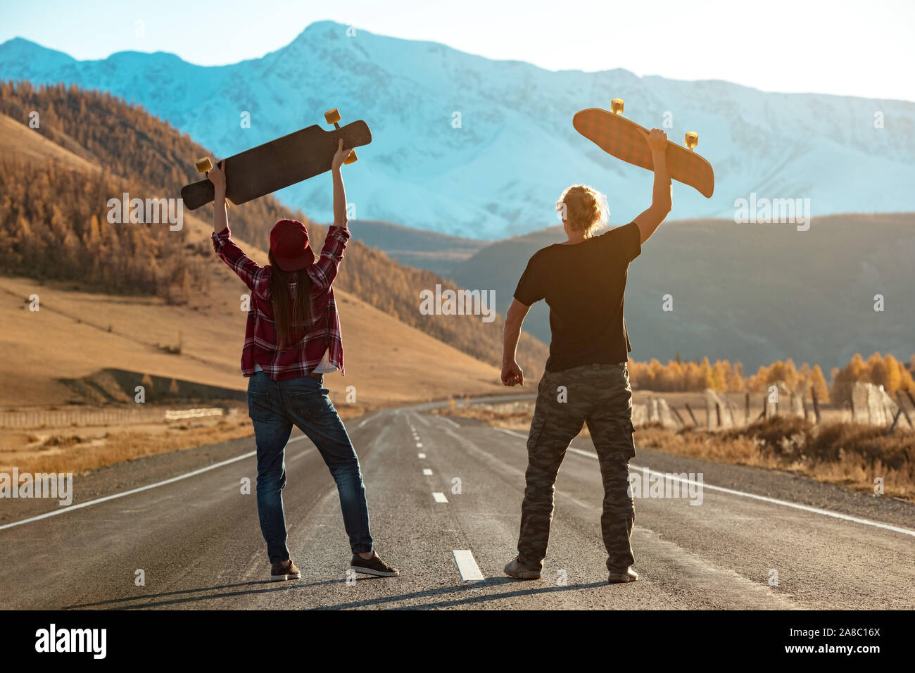 Couple of longboarders stands at straight mountain road with longboard above their heads. Outdoors longboarding concept Stock Photo