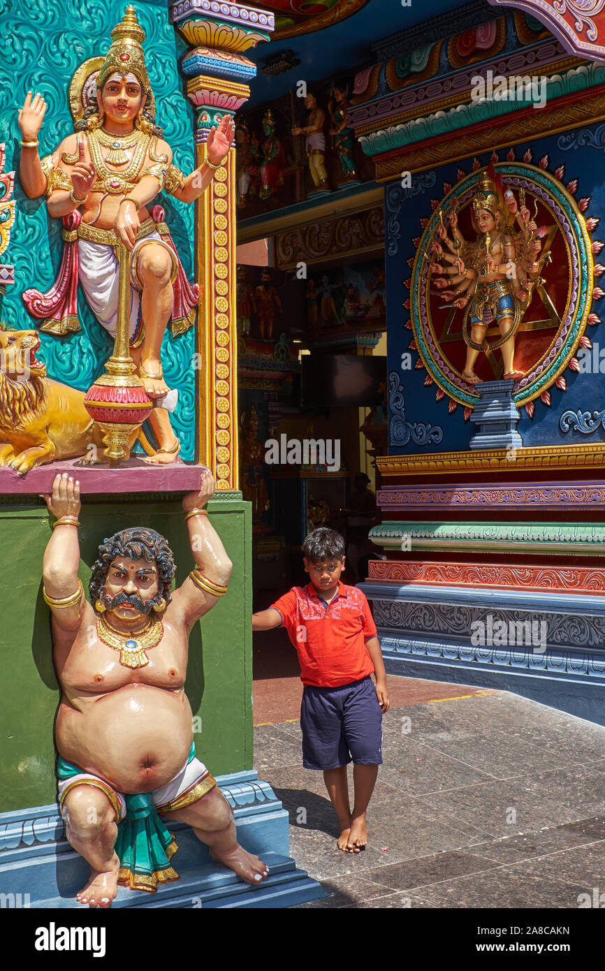 An ethnic Indian boy stands in the entrance to Sri Vadipathira Kaliammam Temple, Serangoon Rd., Singapore, next to a frieze of a Hindu deity Stock Photo