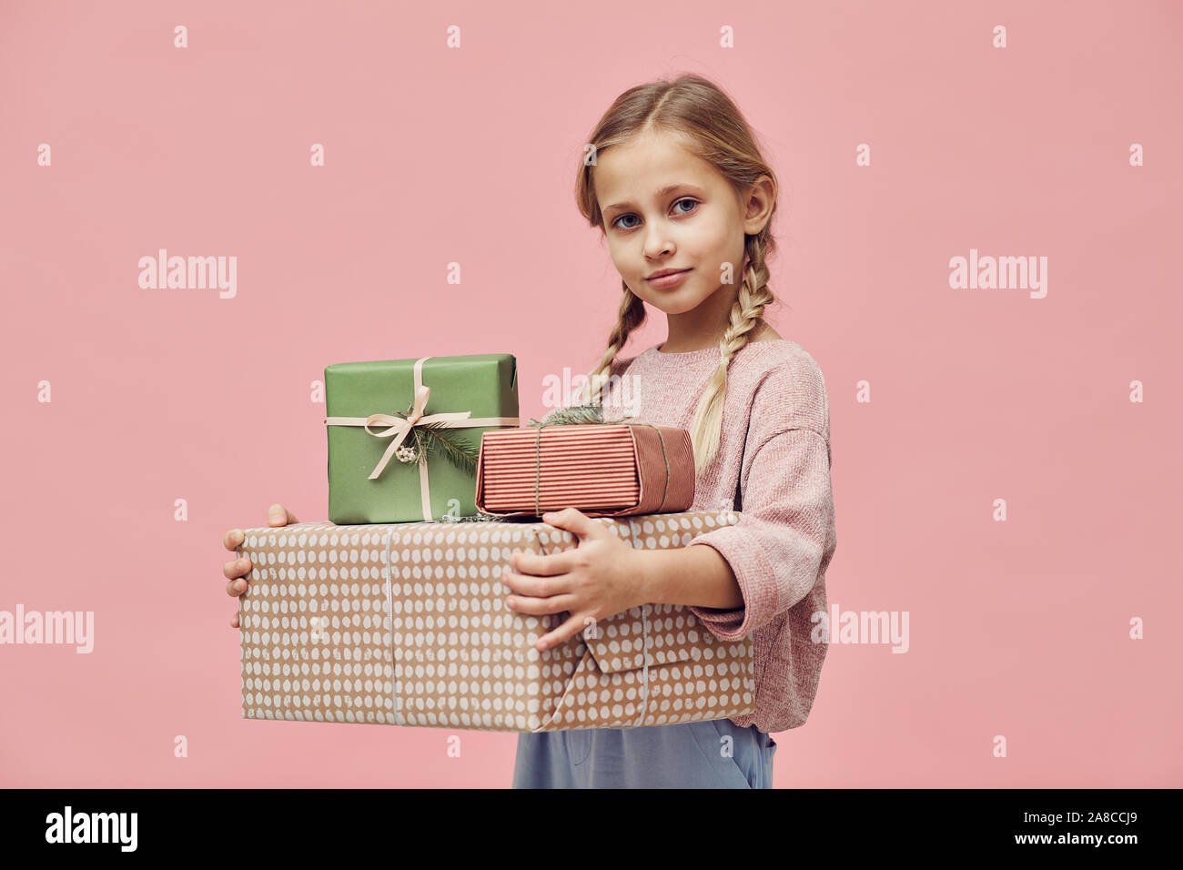 Portrait of little girl with blond hair holding wrapped gift boxes giving her on her birthday and looking at camera over pink background Stock Photo