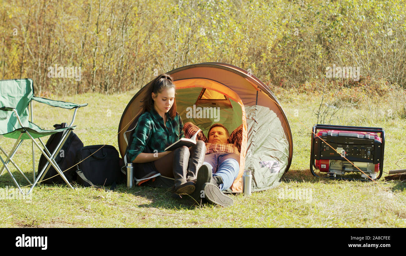 Boyfriend laying down on camping tent while girlfriend's reading a book. Stock Photo