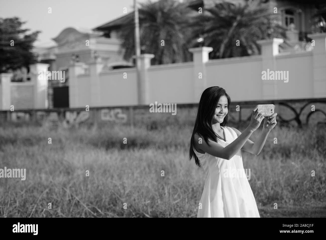 Young happy Asian woman smiling while taking selfie picture with mobile phone against grass field and fancy house Stock Photo