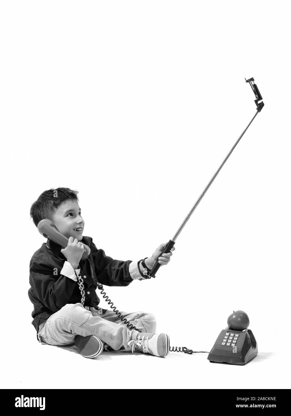 Studio shot of cute happy boy smiling and talking on old telephone while taking selfie with selfie stick Stock Photo