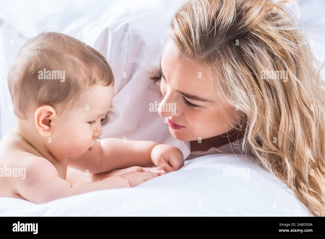 Mother and baby boy son playing on a white bed. Mothers tenderness and kisses of a toddler child. Stock Photo