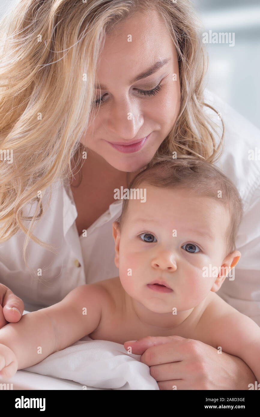 Mother and baby boy son playing on a white bed. Mothers tenderness and kisses of a toddler child. Stock Photo