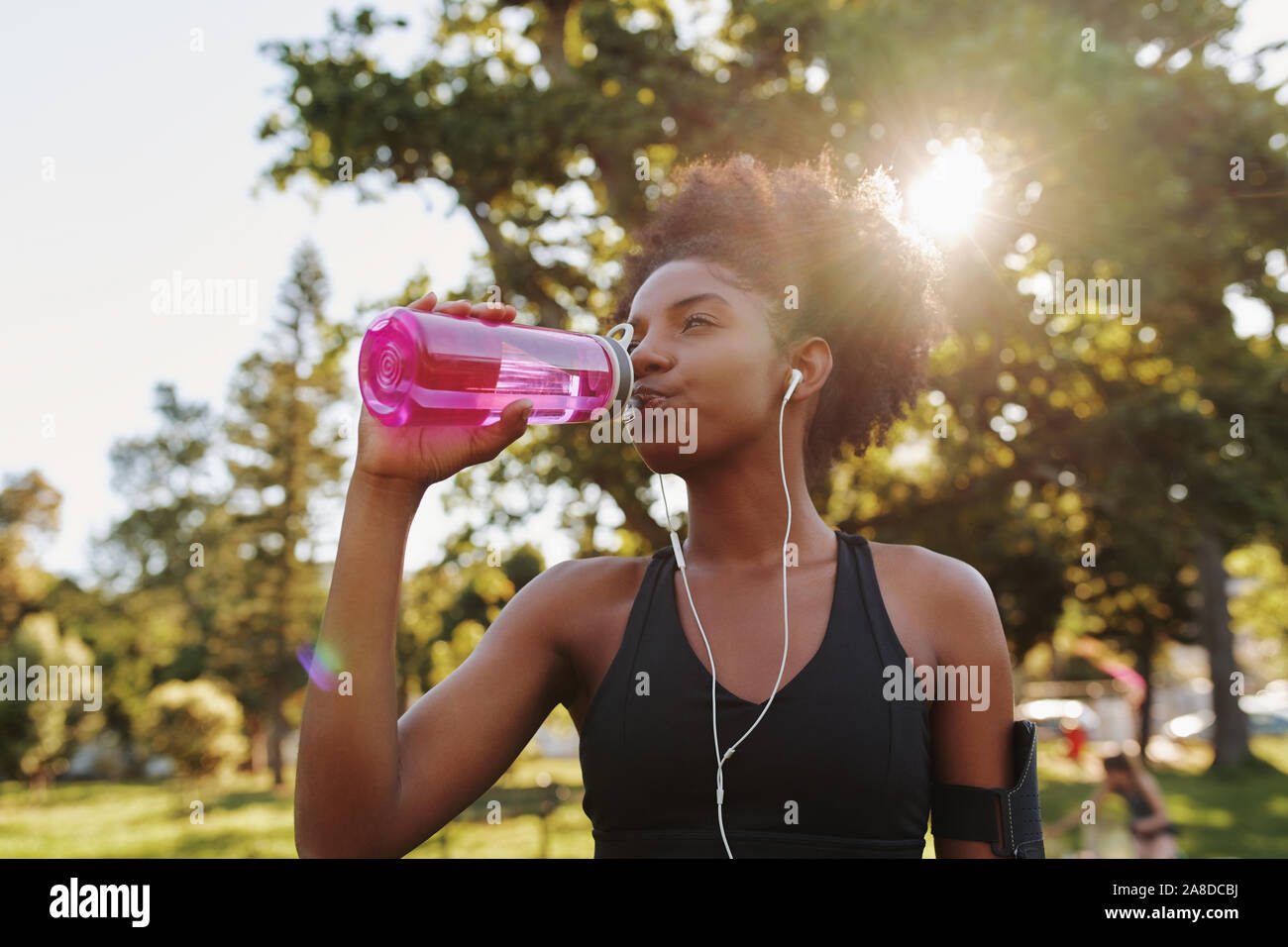 Fitness athlete young african american woman listening to music on earphones drinking water in a reusable water bottle after working out exercising on Stock Photo