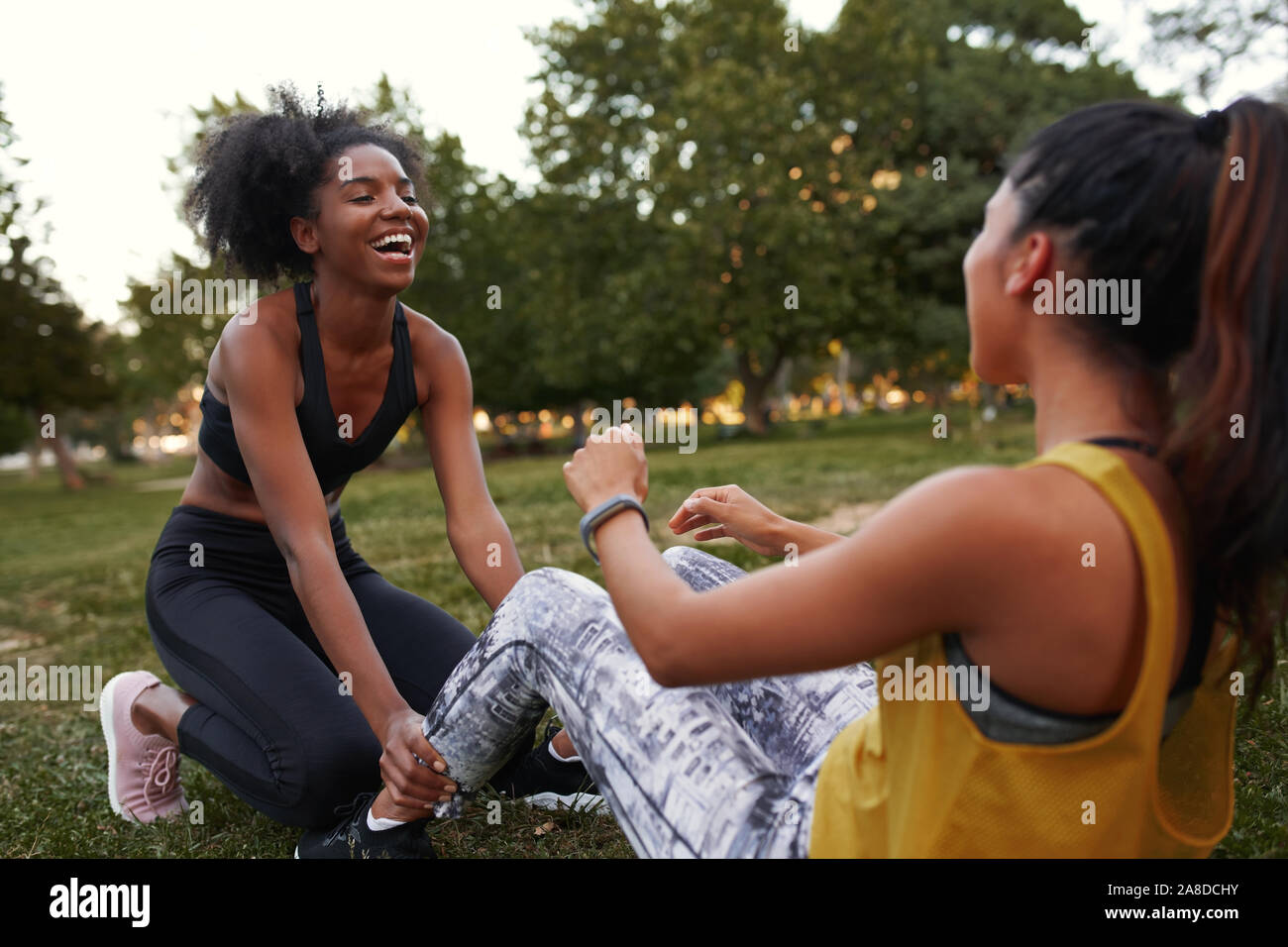 Cheerful young diverse female friends laughing doing ab crunches on green grass in the park - friends laughing together while doing exercise outdoors Stock Photo