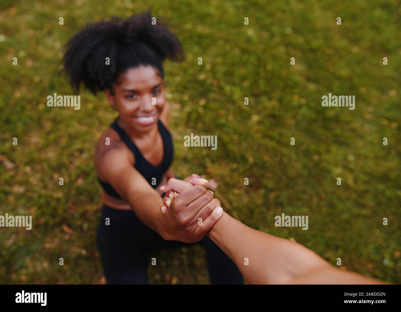 An above view of a person giving helping hand to smiling african american young woman sitting on green grass doing exercise  Stock Photo