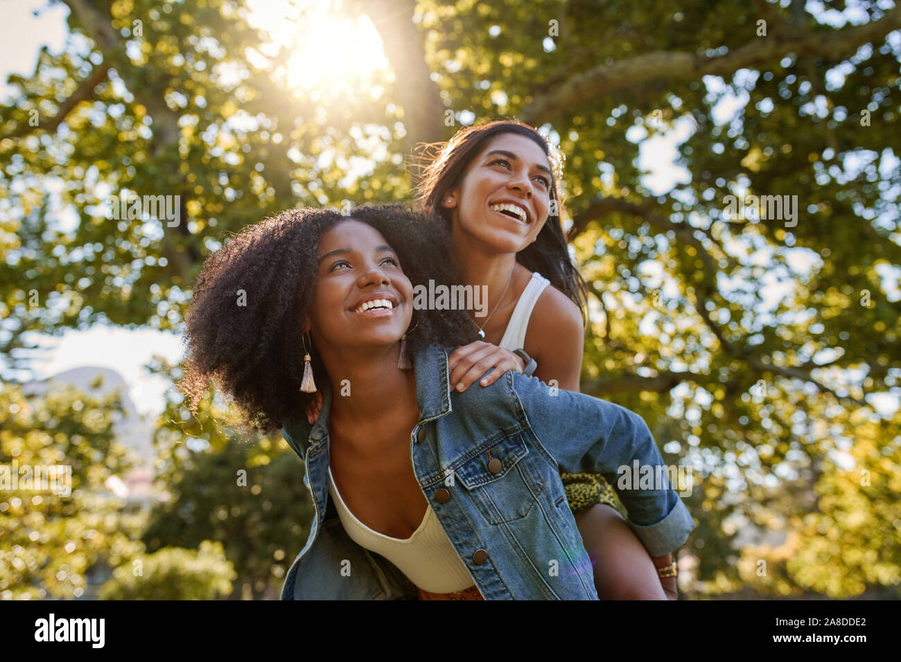 Two diverse multiracial friends having fun laughing and giving a piggyback to each other in the park on a warm summer day Stock Photo