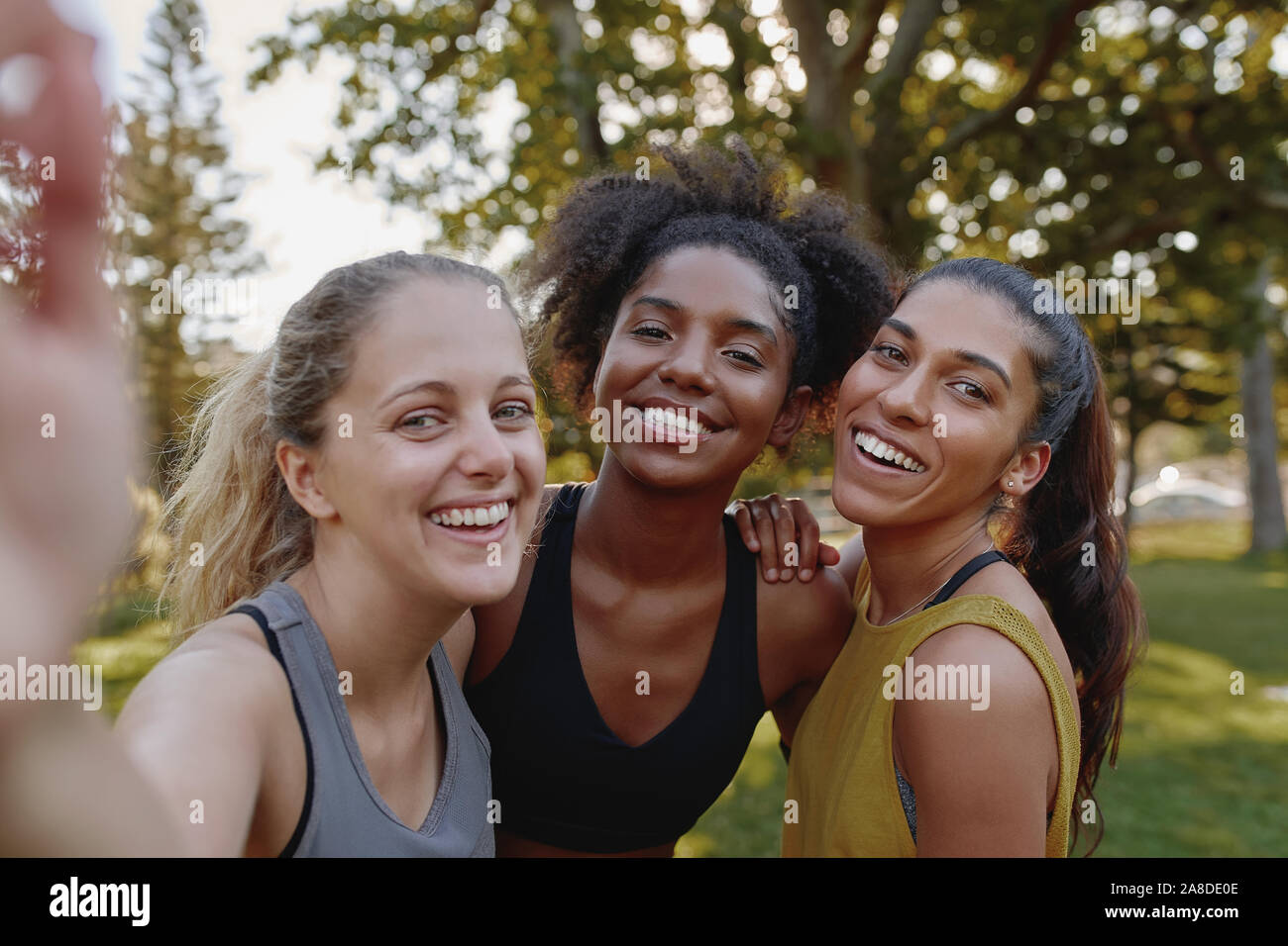 Smiling portrait of diverse female friends looking at camera taking selfie in park - fitness friends taking a selfie after exercising  Stock Photo
