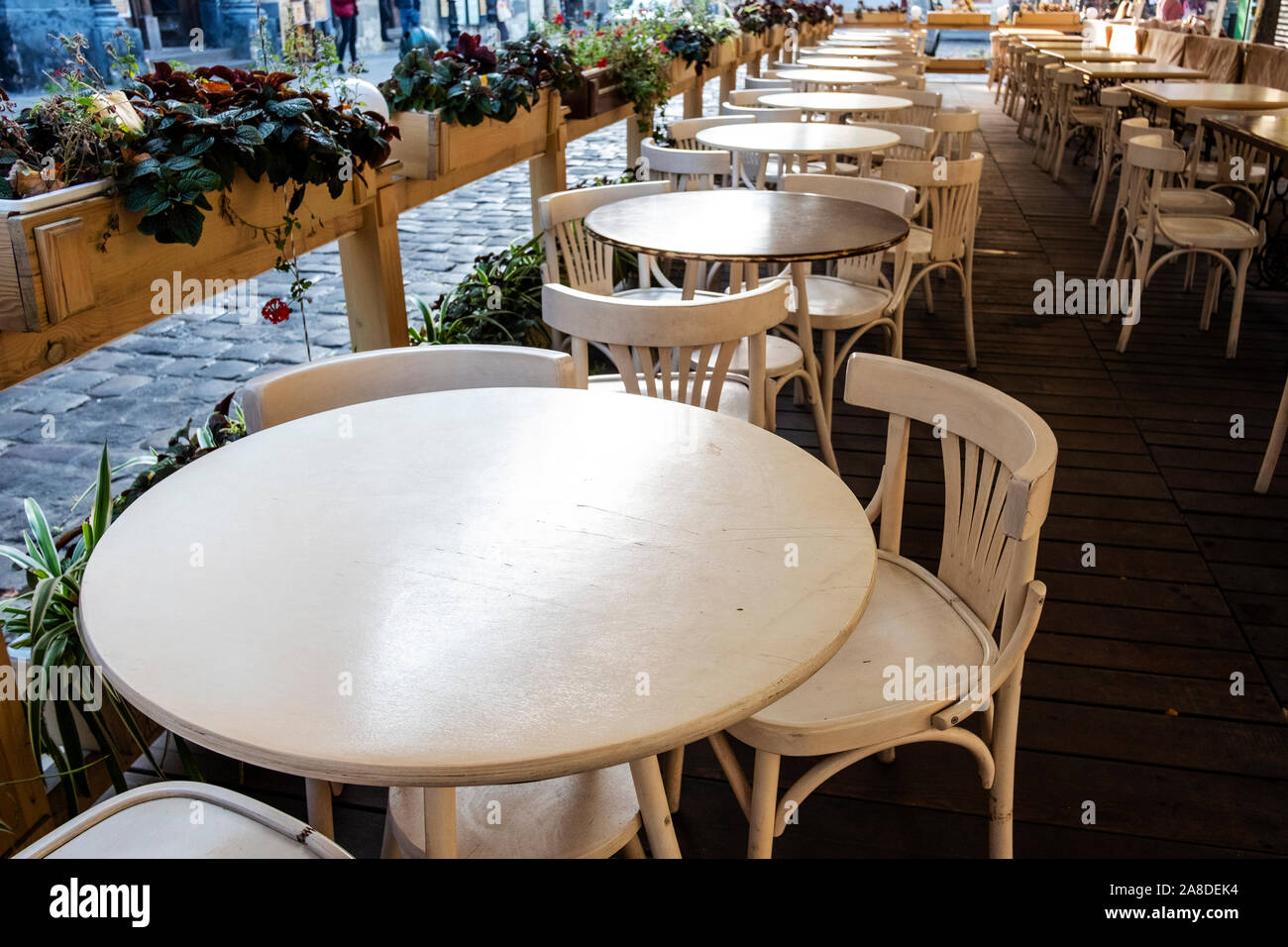 Empty round tables and chairs at an open-air restaurant on a paved street with potted plants in wooden boxes forming a boundary Stock Photo