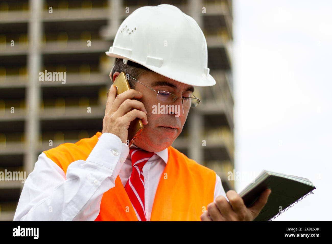 Construction engineer man in shirt and tie with safety helmet and vest works at construction site, talking phone. Concept of people working in industr Stock Photo
