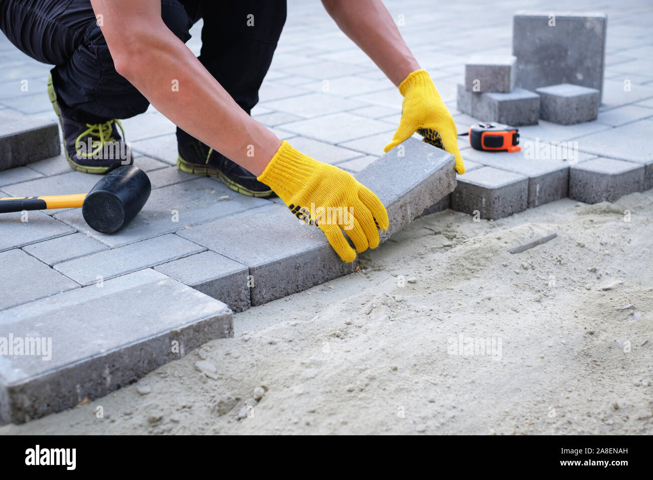 The master in yellow gloves lays paving stones in layers. Garden brick pathway paving by professional paver worker. Laying gray concrete paving slabs Stock Photo