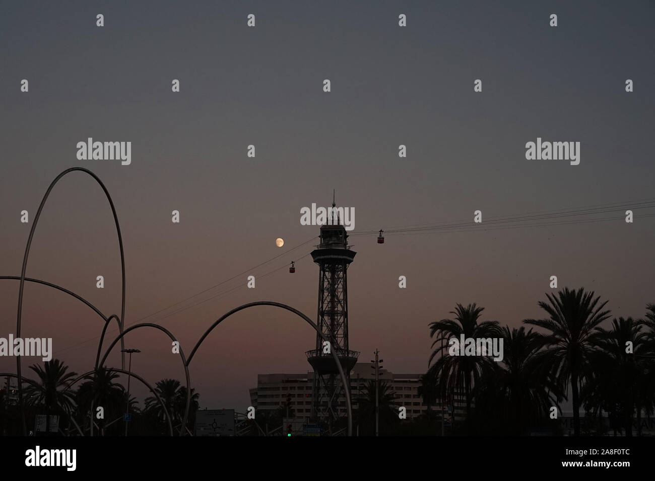 Silhouette of Barcelona cable car tower, palm trees, the moon & structures at sunset Stock Photo