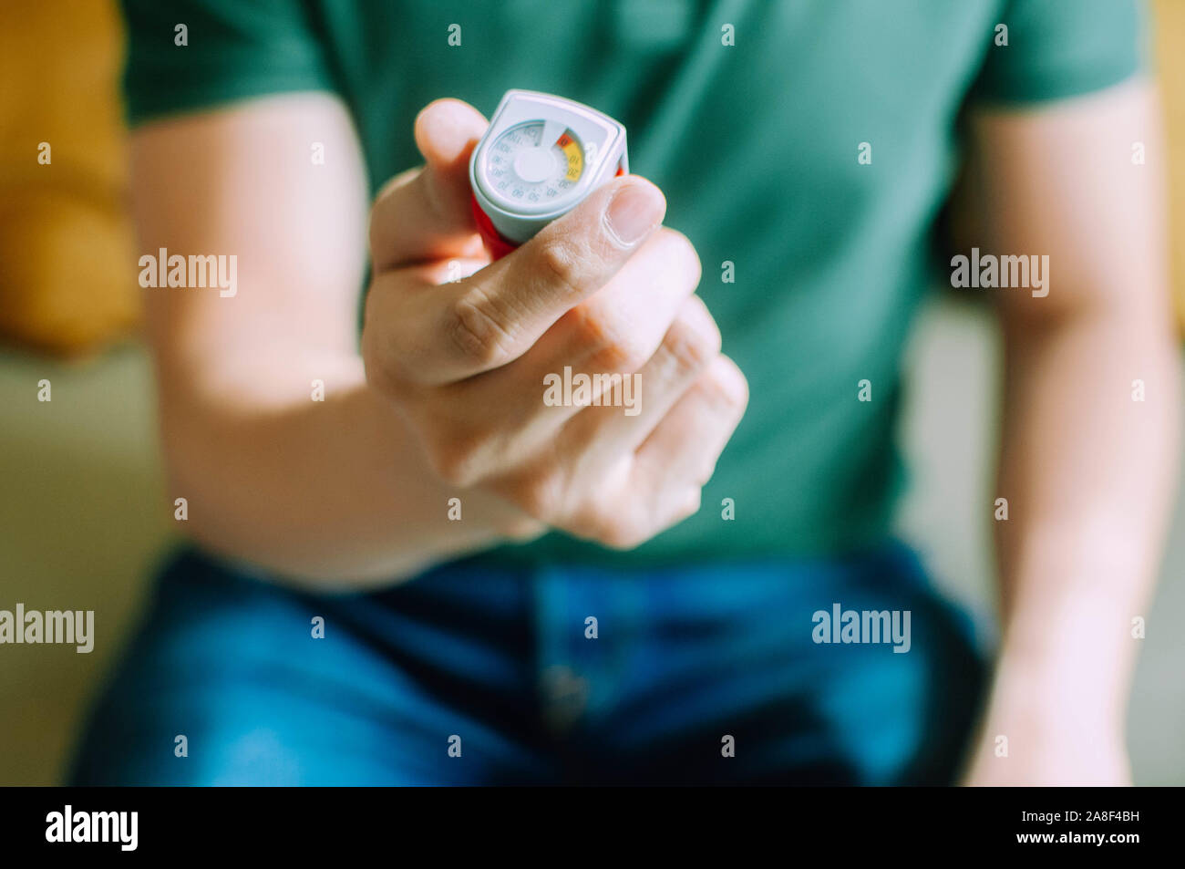 A young man is holding an asthma inhaler device while sitting on a couch Stock Photo