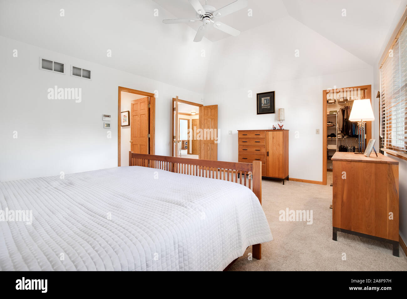 A large master bedroom with tall ceilings, white walls and multiple doorways leading to other rooms. A light brown colored furniture and trim. Stock Photo