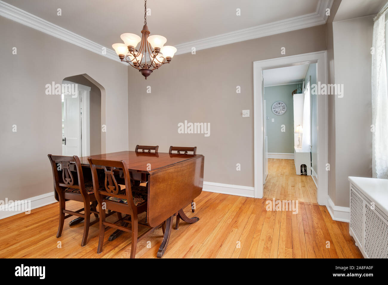 An old plain dining room with a table and chandelier hanging from above. Doorways lead to attached rooms with hardwood floor. Stock Photo