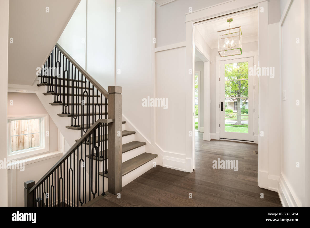 A new construction home with wrought iron railing on the staircase looking out to the front door and green trees and plants outside. Stock Photo