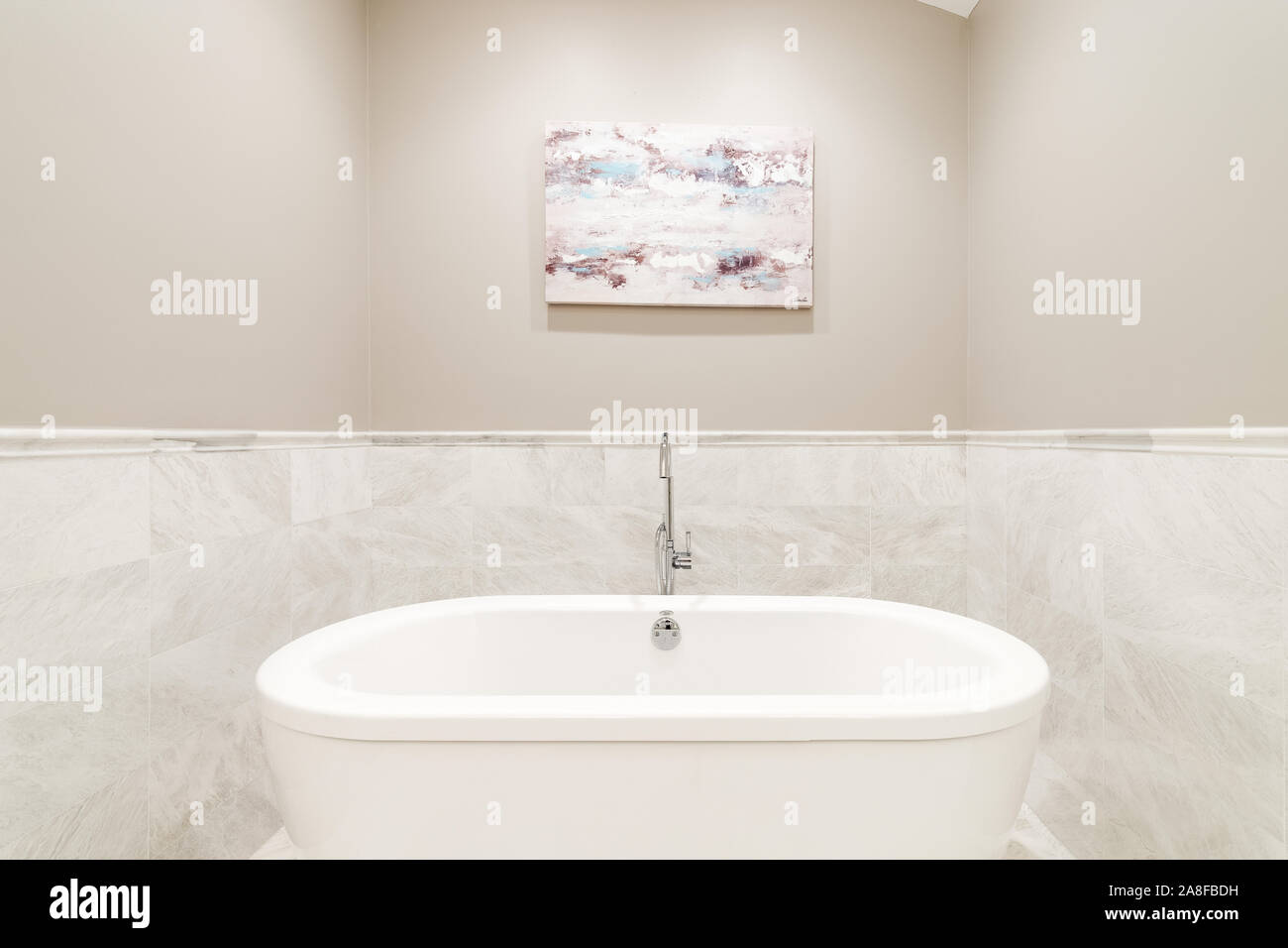 A standalone bathtub sitting in a tan and marble covered nook of a luxurious bathroom with a chrome faucet and a picture hanging above. Stock Photo