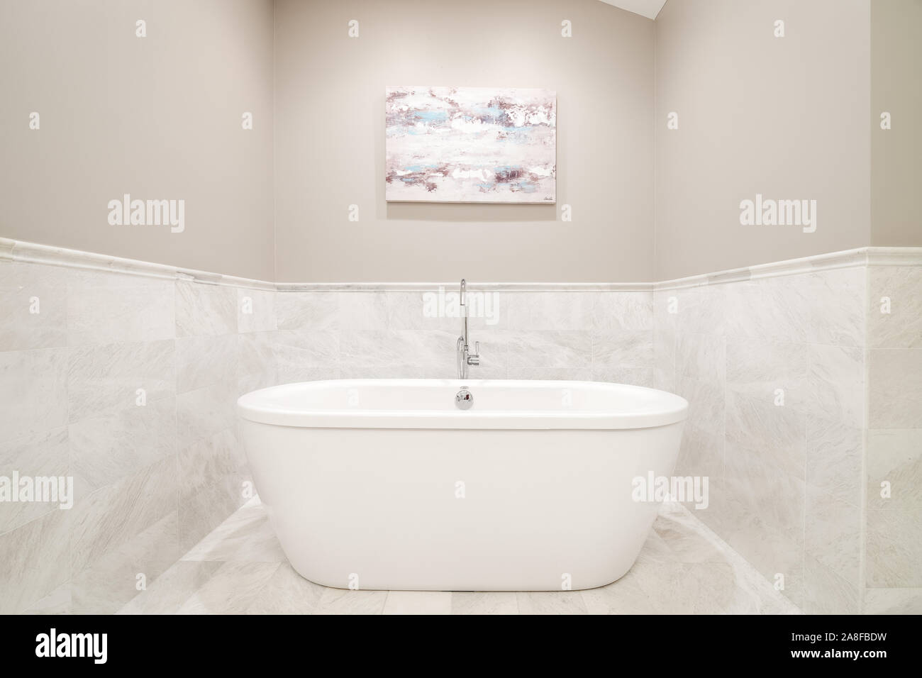 A standalone bathtub sitting in a tan and marble covered nook of a luxurious bathroom with a chrome faucet and a picture hanging above. Stock Photo