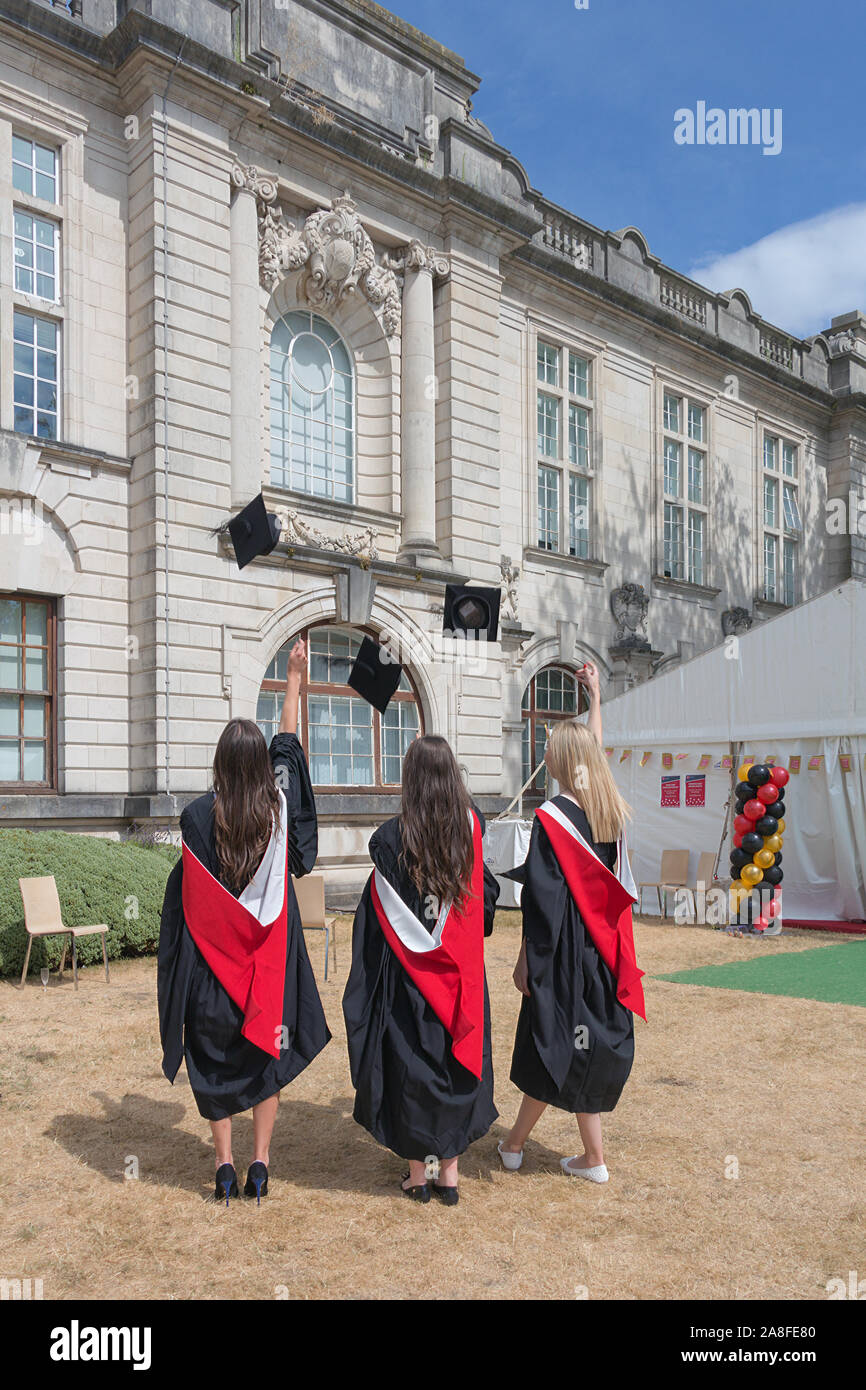 Back side view of three happy girls wearing academic dresses throwing academic caps in yard of Cardiff University, Cardiff, Wales Stock Photo