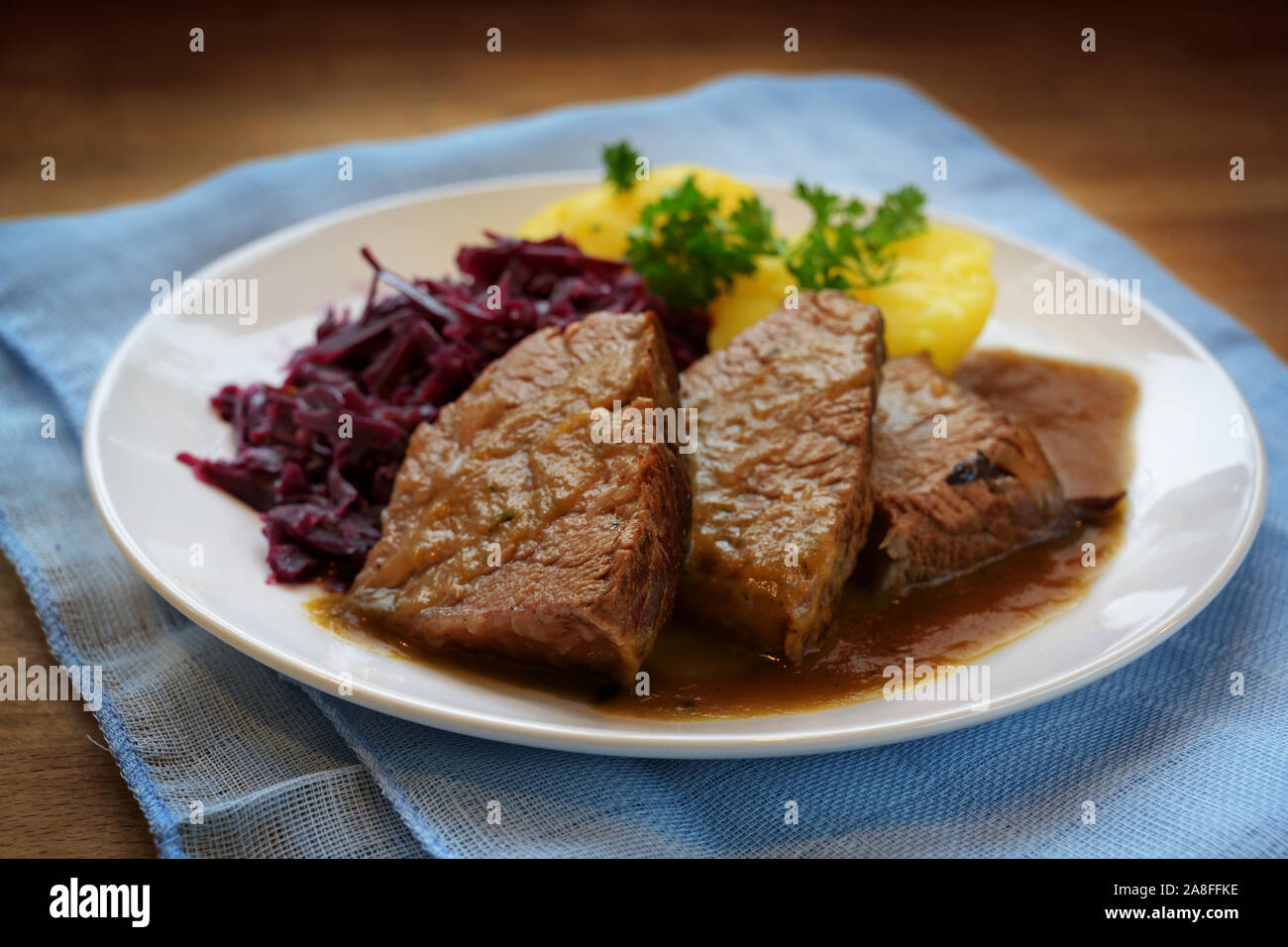 braised beef with sauce, potatoes and red cabbage, served on a white plate and blue napkin on a rustic wooden table, selected focus, very narrow depth Stock Photo