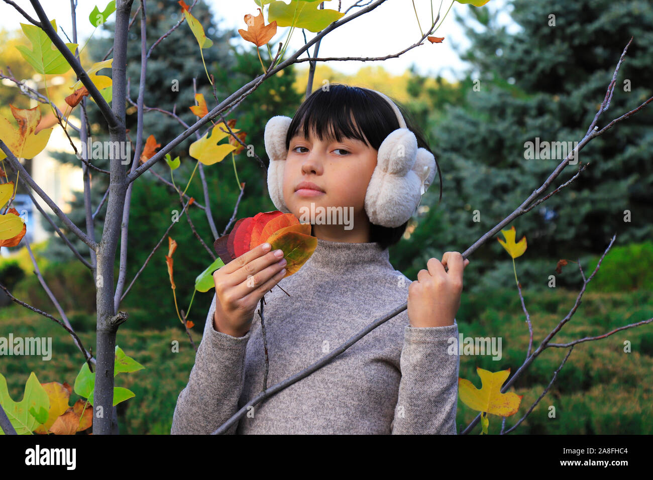 Cute girl teenager looking at the camera near autumn tree. Portrait of girl in white headphones in leaf fall. Dnipro city, Ukraine,12.10.2019 Stock Photo