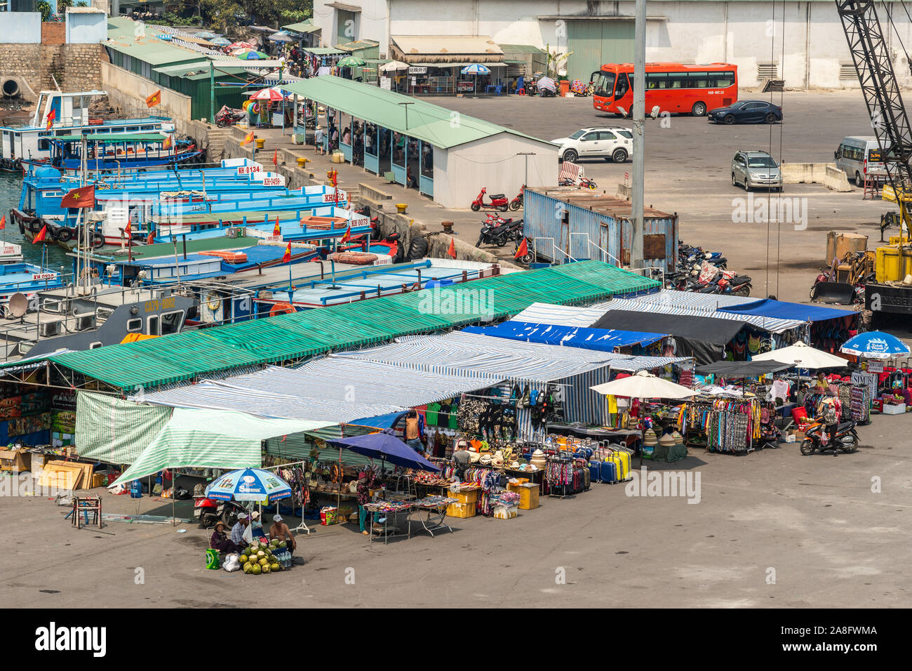 Nha Trang, Vietnam - March 11, 2019: Daily market booths under colored awnings adjacent to ferry landing with several boats at port. People, cars and Stock Photo