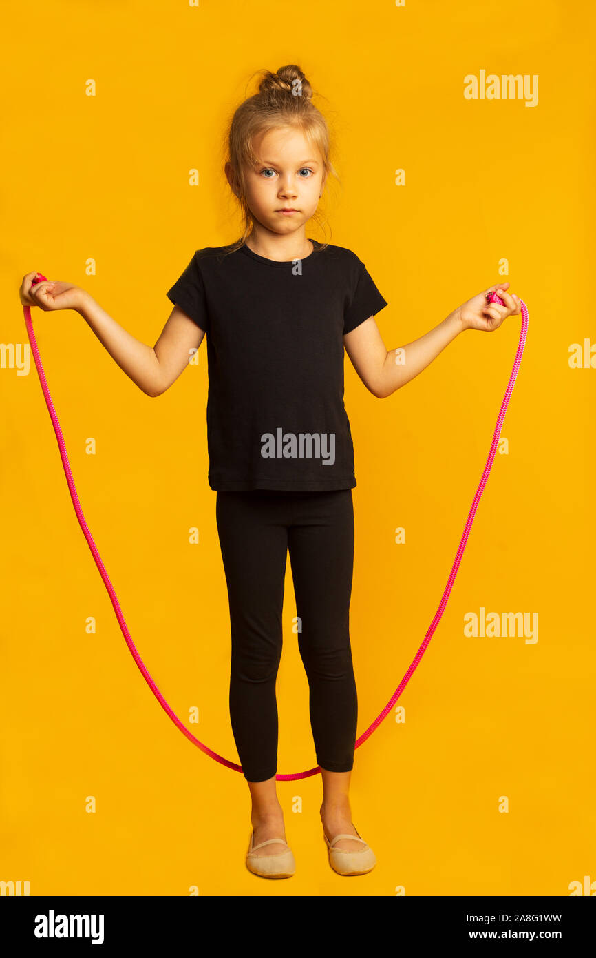 Full length little girl in black leotard jumping on a pink rope against bright yellow background. Stock Photo